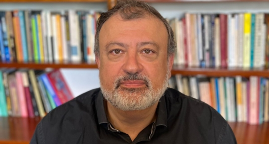 Retrato de Christian Dunker, homem de barba e cabelo grisalhos, com expressão séria, diante de estante de livros