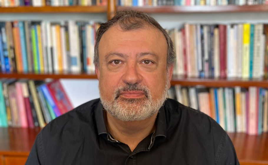 Retrato de Christian Dunker, homem de barba e cabelo grisalhos, com expressão séria, diante de estante de livros