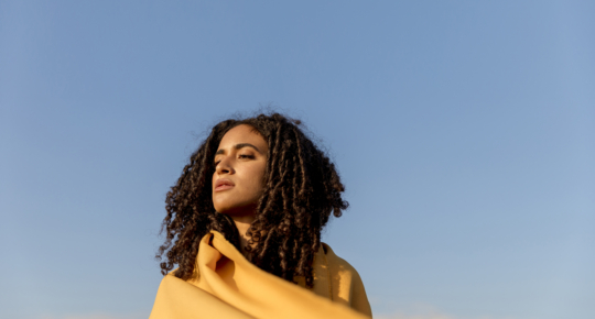 Mulher com cabelos cacheados soltos envolta em um tecido amarelo, olhando para o horizonte sob um céu azul limpo. A imagem transmite sensação de liberdade e contemplação, com luz natural iluminando o rosto e fundo amplo e aberto.
