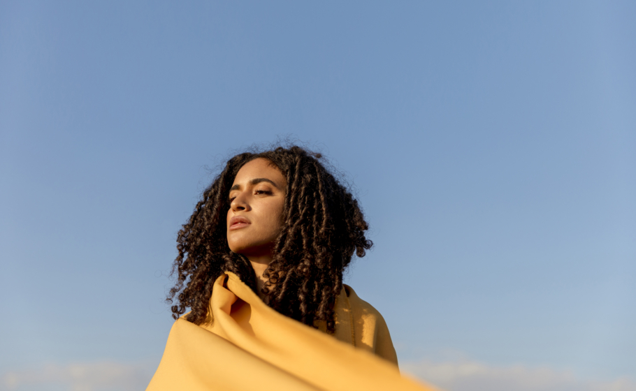 Mulher com cabelos cacheados soltos envolta em um tecido amarelo, olhando para o horizonte sob um céu azul limpo. A imagem transmite sensação de liberdade e contemplação, com luz natural iluminando o rosto e fundo amplo e aberto.