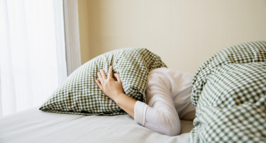 Mulher deitada na cama cobrindo a cabeça com um travesseiro, em posição deitada de lado, demonstrando insônia, indisposição ou tentativa de bloquear ruídos e luz, em ambiente de quarto com lençóis claros.