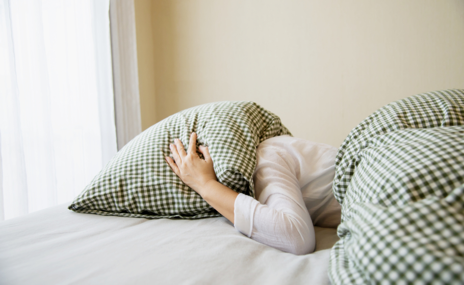 Mulher deitada na cama cobrindo a cabeça com um travesseiro, em posição deitada de lado, demonstrando insônia, indisposição ou tentativa de bloquear ruídos e luz, em ambiente de quarto com lençóis claros.