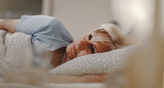 Mulher idosa deitada na cama, apoiando a cabeça na mão e olhando pensativa para o lado, em quarto com roupa de cama clara, transmitindo reflexão, preocupação ou lembranças.