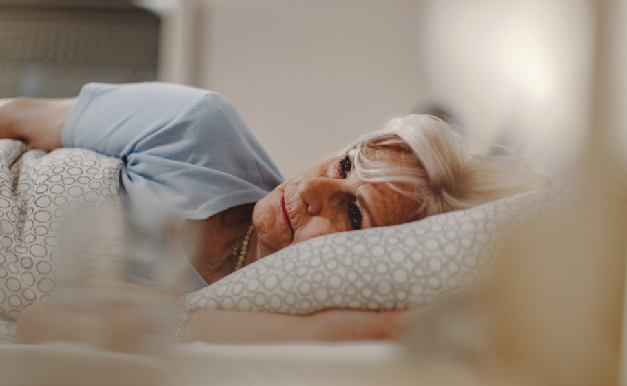 Mulher idosa deitada na cama, apoiando a cabeça na mão e olhando pensativa para o lado, em quarto com roupa de cama clara, transmitindo reflexão, preocupação ou lembranças.