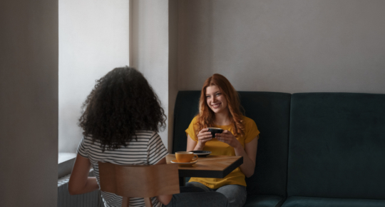 Duas mulheres conversam enquanto tomam café em uma cafeteria. Uma delas sorri segurando uma xícara, e a outra está sentada de costas para a câmera.