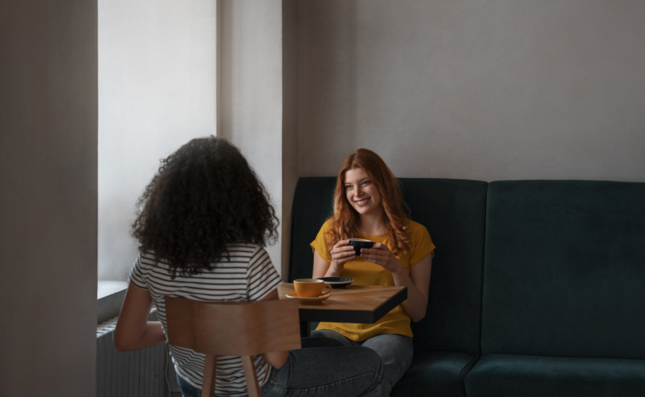 Duas mulheres conversam enquanto tomam café em uma cafeteria. Uma delas sorri segurando uma xícara, e a outra está sentada de costas para a câmera.