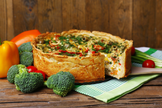 Foto mostra uma quiche de vegetais sobre mesa de madeira, com uma fatia cortada, e rodeada por brócolis, tomates e pimentões
