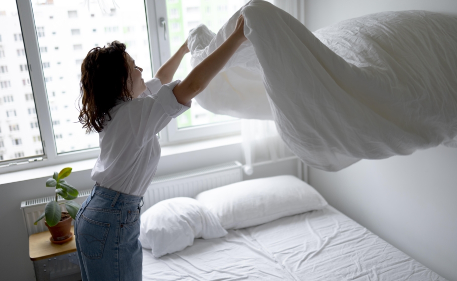 Foto mostra mulher de cabelos castanhos vestindo jeans e blusa branca estendendo edredom na cama diante de uma janela.
