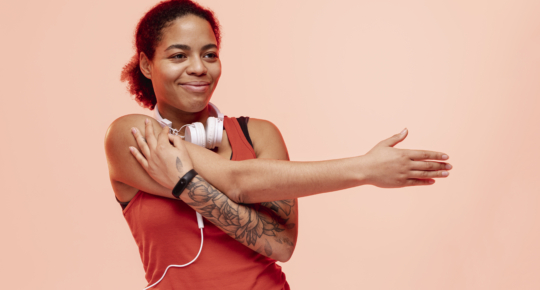 Mulher com cabelos presos, usando blusa de ginástica regata vermelha e fones de ouvido brancos se alonga com uma expressão feliz. O fundo da foto é laranja-claro.