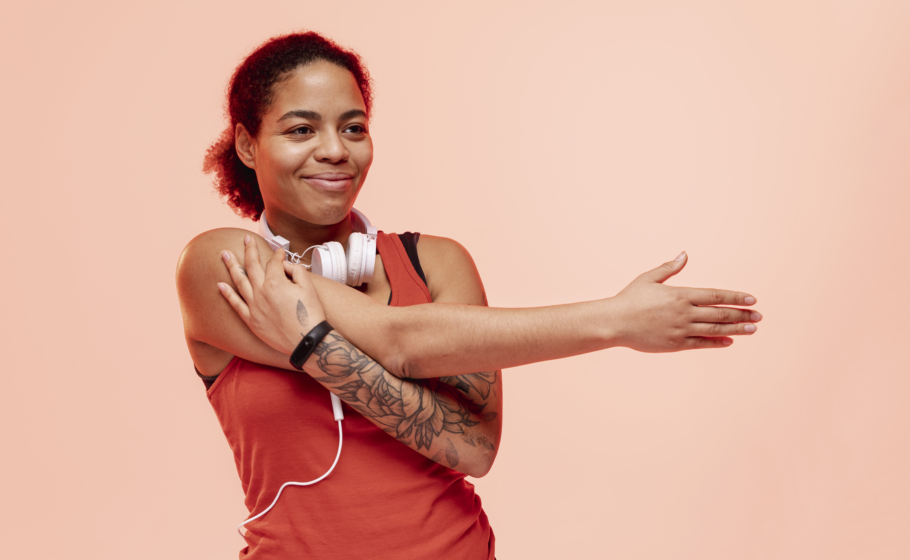 Mulher com cabelos presos, usando blusa de ginástica regata vermelha e fones de ouvido brancos se alonga com uma expressão feliz. O fundo da foto é laranja-claro.