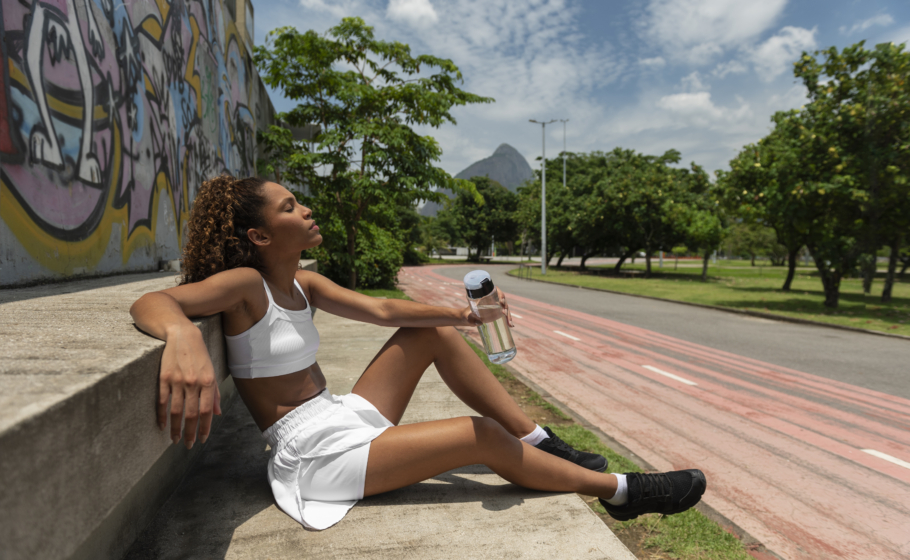 Mulher com roupas esportivas brancas sentada no chão, encostada em um muro com grafite, descansando após o exercício. Ela segura uma garrafa de água e está ao lado de uma pista de corrida ao ar livre, cercada por árvores, com uma montanha ao fundo sob céu parcialmente nublado.