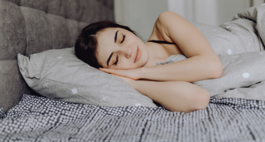 Jovem mulher morena e de cabelos aparentemente presos dormindo na cama, deitada de lado sobre lençóis claros, com expressão serena e relaxada, transmitindo tranquilidade e sono pacífico.