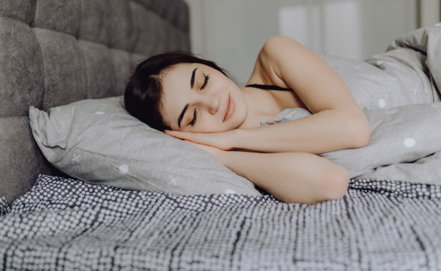 Jovem mulher morena e de cabelos aparentemente presos dormindo na cama, deitada de lado sobre lençóis claros, com expressão serena e relaxada, transmitindo tranquilidade e sono pacífico.