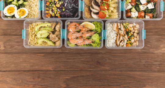 Vista superior de várias marmitas organizadas em potes plásticos transparentes sobre uma mesa de madeira. Cada recipiente contém combinações de alimentos saudáveis, como arroz, feijão, frango grelhado, camarão com limão, macarrão com abacate, ovos cozidos, saladas com tomate e queijo, legumes variados e grãos.