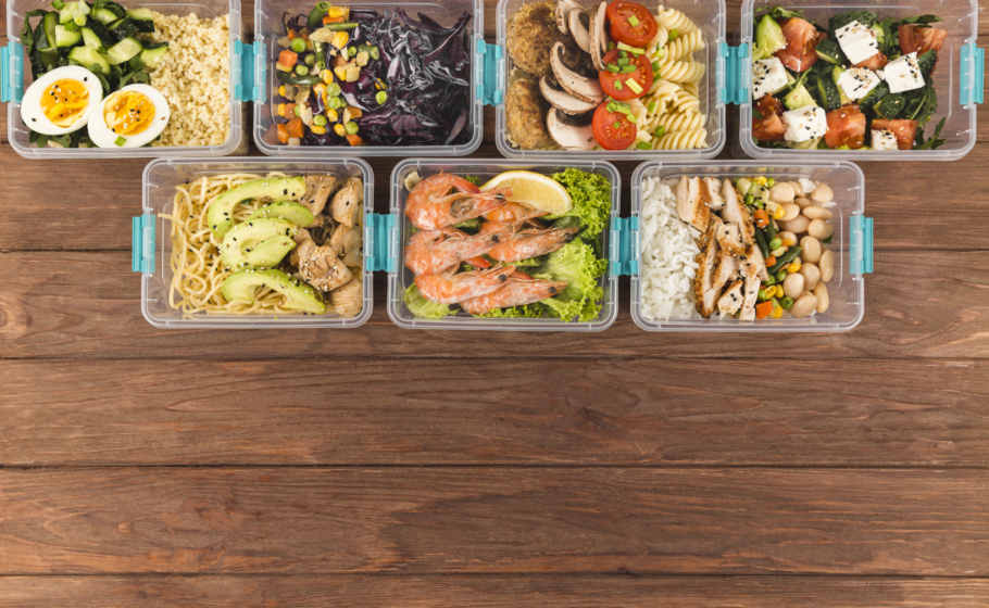 Vista superior de várias marmitas organizadas em potes plásticos transparentes sobre uma mesa de madeira. Cada recipiente contém combinações de alimentos saudáveis, como arroz, feijão, frango grelhado, camarão com limão, macarrão com abacate, ovos cozidos, saladas com tomate e queijo, legumes variados e grãos.