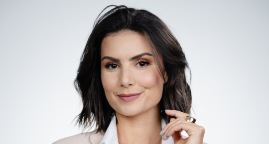 retrato da jornalista Nathalia Arcuri, uma mulher branca, com cabelos pretos na altura do ombro e olhos castanhos, com um braço cruzado sobre o abdômem e vestindo um terninho creme
