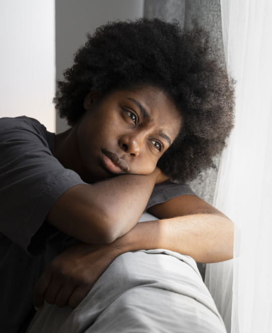 Jovem mulher negra com cabelos black e olhar triste se encosta em um sofá apoiando a cabeça entre os braços
