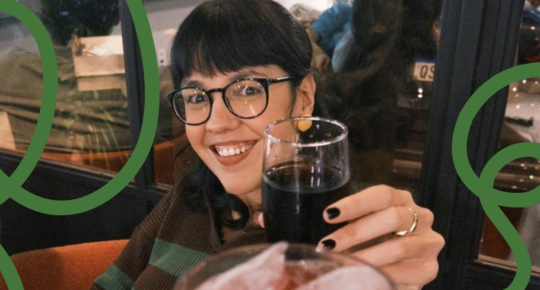 Foto mostra Anna Gouveia, mulher de cabelos lisos, pretos e de franja e óculos, brindando com copo que parece ser de coca cola