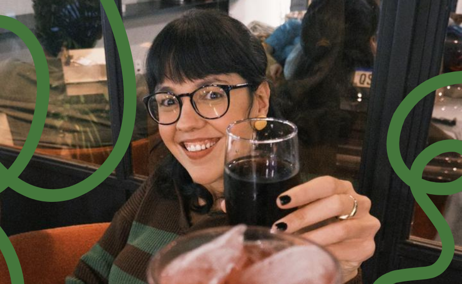 Foto mostra Anna Gouveia, mulher de cabelos lisos, pretos e de franja e óculos, brindando com copo que parece ser de coca cola