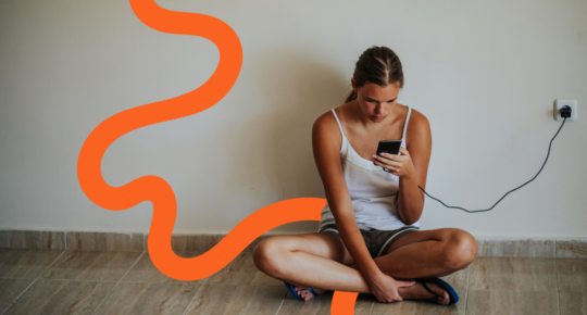 Em arte, jovem usando camisa regata branca usa o celular no chão, enquanto ele carrega na tomada
