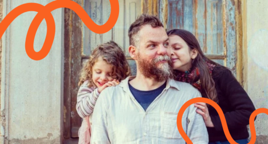 Arte mostra Marcos Piangers, um homem branco e com barba, ao lado das duas filhas, duas crianãs bancas abraçadas ao pai